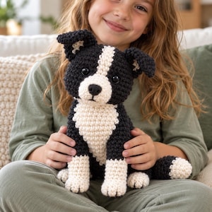 May include: A handmade crochet stuffed animal in the likeness of a dog. The plush toy features a black and cream color scheme with a white stripe running down the center of its face, black ears, dark plastic eyes, and a black stitched nose.