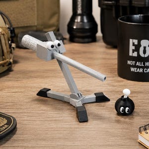 Op de afbeelding: Een grijze plastic gearticuleerde robotfiguur met wiebeloogjes en een lange arm, naast een klein zwart bolvormig bom-speeltje met een witte lont en wiebeloogjes. Een zwarte mok bevat de tekst EOD NOT ALL HEROES WEAR CAPES.