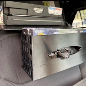 May include: A black rectangular dispenser box with a horizontal oval opening, featuring a side panel with a pattern of oval cutouts. The box contains a stack of dark gloves, with one glove partially pulled through the front opening for easy access.