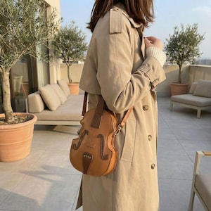 Peut inclure: Sac bandoulière marron en forme de violon. Le sac présente un design détaillé imitant un violon, avec un accent marron plus foncé en haut et en bas. Une personne porte le sac, vêtue d'un trench-coat beige.