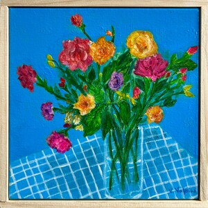 May include: An original painting of a colorful floral bouquet in a clear vase. The flowers are in shades of pink, orange, yellow, and purple, set against a blue background and a blue and white checkered tablecloth. The painting is framed in a light wood frame.