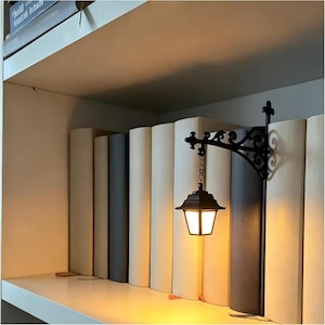 May include: A black metal wall-mounted lantern with an ornate scrollwork bracket, suspended by a small chain. The lantern features a glowing, warm-toned light source enclosed within a geometric frame, positioned between a row of upright books.