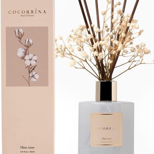 May include: A Cocorrína reed diffuser with a light gray glass bottle, a gold collar, and dark brown reeds topped with dried white baby's breath flowers. The matching beige rectangular box features a cotton branch illustration and the text Clean Linen.