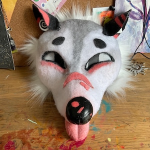 May include: A handcrafted animal mask featuring white and gray faux fur, black mesh eye coverings, and pointed ears with black and red accents. The face has a black nose with a pink spot, pink markings under the eyes, and a pink tongue extending downward.