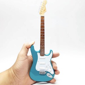 Puede incluir: Una guitarra el&eacute;ctrica en miniatura de color turquesa con un golpeador blanco y herrajes plateados. La guitarra tiene m&aacute;stil y clavijero de madera. El instrumento se sostiene en una mano sobre un fondo blanco.