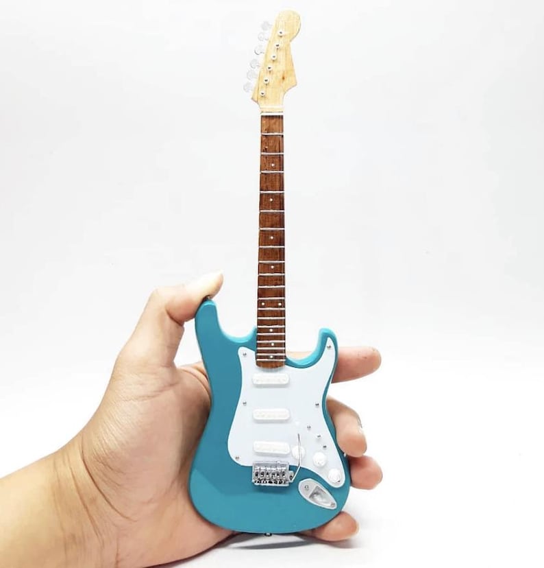 Puede incluir: Una guitarra el&eacute;ctrica en miniatura de color turquesa con un golpeador blanco y herrajes plateados. La guitarra tiene m&aacute;stil y clavijero de madera. El instrumento se sostiene en una mano sobre un fondo blanco.