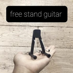 Puede incluir: Un peque&ntilde;o soporte negro para guitarra con un dise&ntilde;o de gancho de metal, sostenido en una mano. El soporte es compacto. El texto "free stand guitar" est&aacute; en un rect&aacute;ngulo negro encima del soporte.