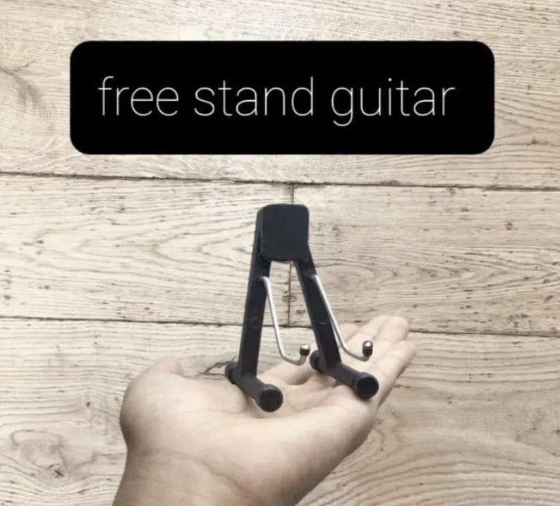 Puede incluir: Un peque&ntilde;o soporte negro para guitarra con un dise&ntilde;o de gancho de metal, sostenido en una mano. El soporte es compacto. El texto "free stand guitar" est&aacute; en un rect&aacute;ngulo negro encima del soporte.