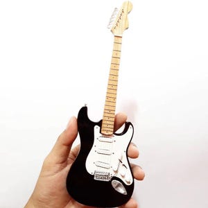 Puede incluir: Miniatura de guitarra el&eacute;ctrica en blanco y negro. La guitarra tiene un cuerpo negro, un golpeador blanco y un m&aacute;stil y clavijero de color claro. La guitarra tiene seis cuerdas y herrajes plateados. El instrumento se sostiene en una mano.