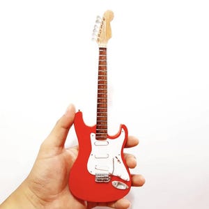 Puede incluir: Una guitarra el&eacute;ctrica roja en miniatura con un golpeador blanco y herrajes plateados. La guitarra tiene m&aacute;stil y clavijero de madera. El instrumento se sostiene sobre un fondo blanco.