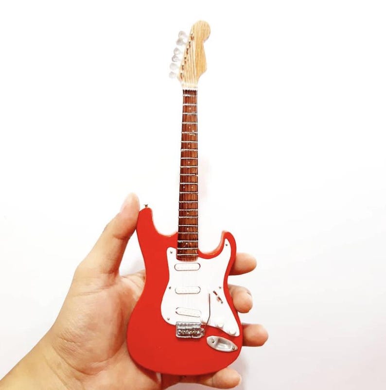 Puede incluir: Una guitarra el&eacute;ctrica roja en miniatura con un golpeador blanco y herrajes plateados. La guitarra tiene m&aacute;stil y clavijero de madera. El instrumento se sostiene sobre un fondo blanco.