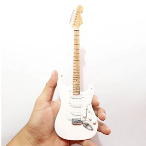 Puede incluir: Una guitarra el&eacute;ctrica blanca en miniatura con m&aacute;stil y clavijero de madera clara. La guitarra tiene herrajes plateados, incluido el puente, las clavijas y las pastillas. El diapas&oacute;n tiene peque&ntilde;os marcadores de traste oscuros. La guitarra se sostiene en una mano sobre un fondo blanco.