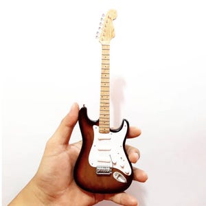 Puede incluir: Miniatura de guitarra el&eacute;ctrica con acabado sunburst, golpeador blanco y herrajes cromados. La guitarra tiene m&aacute;stil y clavijero de color claro. El instrumento se sostiene sobre un fondo blanco.