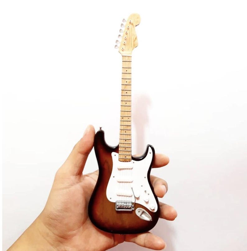 Puede incluir: Miniatura de guitarra el&eacute;ctrica con acabado sunburst, golpeador blanco y herrajes cromados. La guitarra tiene m&aacute;stil y clavijero de color claro. El instrumento se sostiene sobre un fondo blanco.