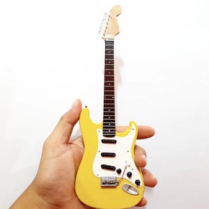 Puede incluir: Una guitarra el&eacute;ctrica amarilla en miniatura con un golpeador blanco y pastillas negras. La guitarra tiene m&aacute;stil y clavijero de madera, y herrajes plateados. El instrumento se sostiene en una mano sobre un fondo blanco.