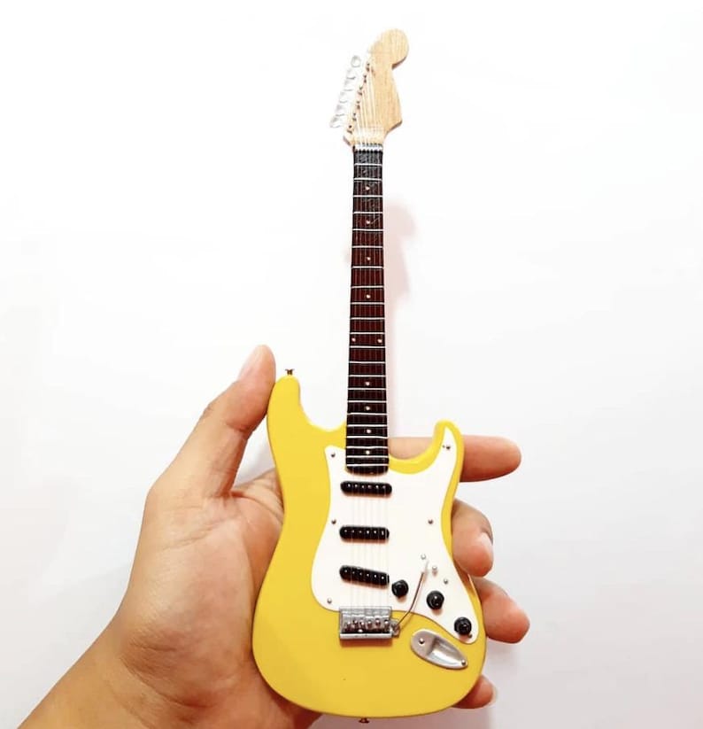 Puede incluir: Una guitarra el&eacute;ctrica amarilla en miniatura con un golpeador blanco y pastillas negras. La guitarra tiene m&aacute;stil y clavijero de madera, y herrajes plateados. El instrumento se sostiene en una mano sobre un fondo blanco.