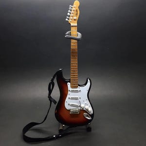 Puede incluir: Una guitarra el&eacute;ctrica en miniatura con acabado sunburst, golpeador blanco y correa negra. La guitarra se exhibe sobre un soporte contra un fondo oscuro. El logotipo de Fender est&aacute; en la pala.