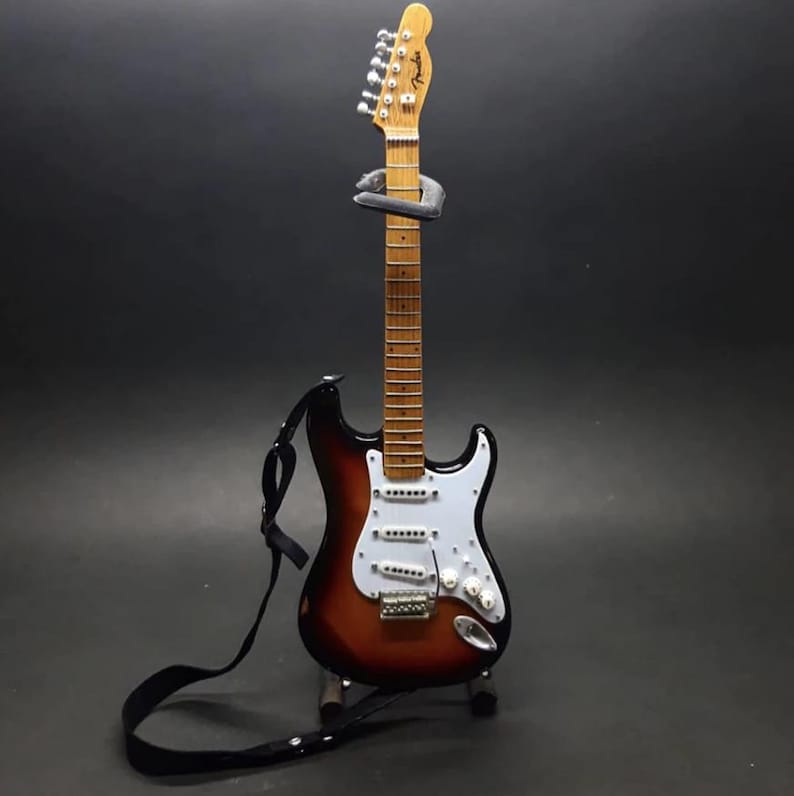Puede incluir: Una guitarra el&eacute;ctrica en miniatura con acabado sunburst, golpeador blanco y correa negra. La guitarra se exhibe sobre un soporte contra un fondo oscuro. El logotipo de Fender est&aacute; en la pala.