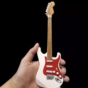 Puede incluir: Una guitarra el&eacute;ctrica en miniatura blanca y roja con m&aacute;stil y clavijero de madera. La guitarra tiene herrajes plateados y se sostiene sobre un fondo negro. La guitarra es una r&eacute;plica en miniatura de un instrumento de tama&ntilde;o completo.