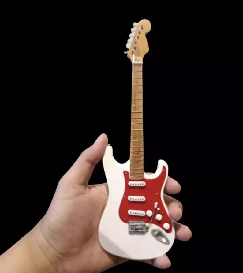 Puede incluir: Una guitarra el&eacute;ctrica en miniatura blanca y roja con m&aacute;stil y clavijero de madera. La guitarra tiene herrajes plateados y se sostiene sobre un fondo negro. La guitarra es una r&eacute;plica en miniatura de un instrumento de tama&ntilde;o completo.