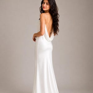 May include: A floor-length white satin gown featuring a deep cowl back, thin spaghetti straps, and a fitted bodice that flows into a straight skirt. The fabric has a smooth, reflective finish, and the design includes a low-cut back neckline.