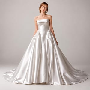 May include: A floor-length white satin ball gown featuring a strapless bodice with a straight neckline, a fitted waistline, and a voluminous skirt with deep pleats that extend into a long train, creating a smooth and lustrous appearance.