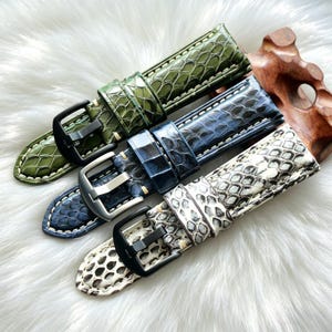 Könnte beinhalten: Vier Uhrenarmbänder in Grün-, Blau- und Weißtönen mit Schlangenmuster. Jedes Armband hat eine Schnalle und Kontrastnähte. Die Armbänder werden auf einer weißen, flauschigen Oberfläche präsentiert.