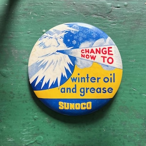 May include: A blue and yellow round button with a cartoon image of a winter figure blowing snow. The button says "Change Now To Winter Oil and Grease Sunoco".