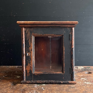 May include: A small, black wooden cabinet with a glass door and a distressed finish. The cabinet has a simple design with a single shelf inside.