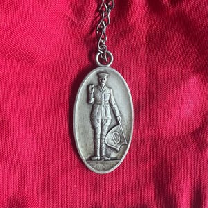 Op de afbeelding: Een zilveren ovale hanger met een ketting. De hanger heeft een reliëfbeeld van een persoon in militair uniform die een vlag vasthoudt.