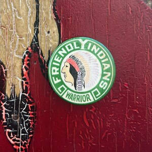 May include: Green button with the words "Friendly Indian Warrior" in a circle. The button features a cartoon image of a Native American wearing a headdress.