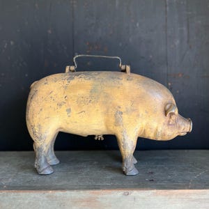 May include: A vintage, yellow painted, wooden piggy bank with a metal handle and a metal latch on the side. The piggy bank is standing on a wooden surface.