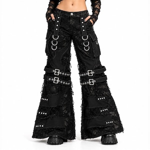 May include: Black wide-leg pants featuring distressed lace overlays, multiple cargo pockets, and silver-tone metal hardware. The design includes adjustable buckled straps across the thighs, vertical straps with D-rings at the waist, and studded panels.