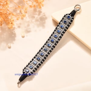 Puede incluir: Una pulsera de cordón negro con una serie de piedras preciosas azules ovaladas engastadas en marcos de metal plateado, rodeadas de pequeñas piedras pavé transparentes. La pulsera incluye un cierre de mosquetón y el texto uniquejewellery925 en azul.