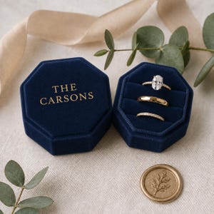 May include: A navy blue velvet octagonal ring box with the text THE CARSONS in gold, containing three gold rings with a central oval diamond. A gold wax seal with a leaf design sits nearby, accompanied by a light beige ribbon and green leaves.