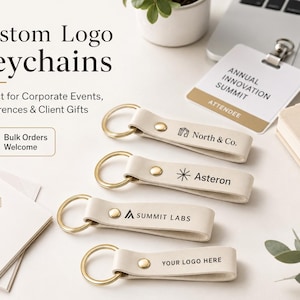 May include: Four off-white leather keychains with gold-tone metal rings and rivets. Each keychain features a unique black printed logo: North & Co., Asteron, Summit Labs, and the placeholder text YOUR LOGO HERE. The keychains are arranged horizontally.