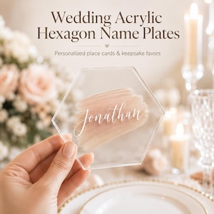 Op de afbeelding: Een doorzichtig zeshoekig naamplaatje van acryl met een handgeschilderde poederroze penseelstreek en de naam Jonathan in witte cursieve letters. De tekst Wedding Acrylic Hexagon Name Plates en Personalized place cards & keepsake favors is zichtbaar.