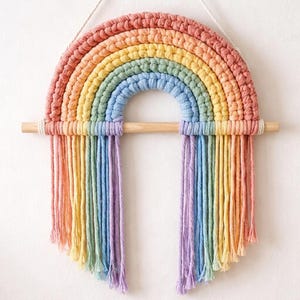Op de afbeelding: Een wandhanger met een regenboogontwerp gemaakt van dik, gevlochten katoenen koord in de kleuren koraal, geel, saliegroen en lichtblauw. De regenboog is bevestigd aan een horizontale houten stok met lange, losse franjes die naar beneden hangen.