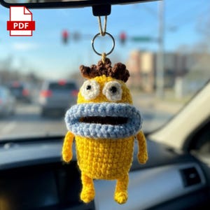 Puede incluir: Un adorno de personaje tejido a ganchillo en color amarillo que cuelga de una anilla metálica. La figura tiene pelo castaño, grandes ojos blancos con pupilas negras y una boca prominente de color azul claro. Cuerpo amarillo con extremidades.