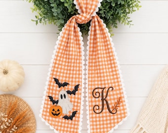May include: An orange and white gingham fabric door hanger featuring a scalloped white edge. One side displays an embroidered white ghost, a small orange pumpkin, and black bats. The other side features a large, dark brown letter K.