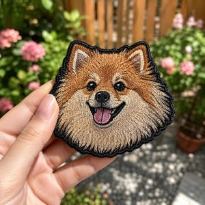 May include: Embroidered patch featuring a smiling Pomeranian dog with brown and tan fur, a pink tongue, and black eyes. The patch has a black border and is held up against a blurred background of flowers and greenery.