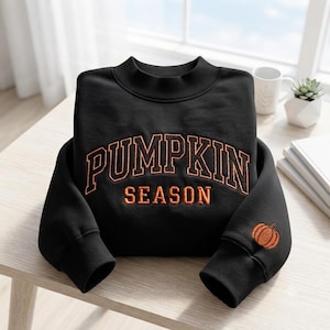 May include: A black long-sleeve sweatshirt with a mock neck collar. The front features the text PUMPKIN SEASON embroidered in orange with a dark outline. A small orange pumpkin icon is embroidered on the lower part of the right sleeve.