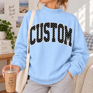 May include: A light blue long-sleeve sweatshirt with a mock neck collar. The front features the word CUSTOM in large, arched, black block letters with a white polka dot pattern. The sweatshirt is paired with a plain white shoulder bag strap.