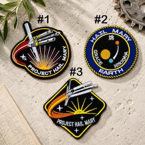 May include: Three embroidered patches with space-themed designs. The patches are black with colorful accents, featuring rockets, sunbursts, and the text "PROJECT HAIL MARY" and "HAIL MARY EARTH". The patches are numbered #1, #2, and #3.
