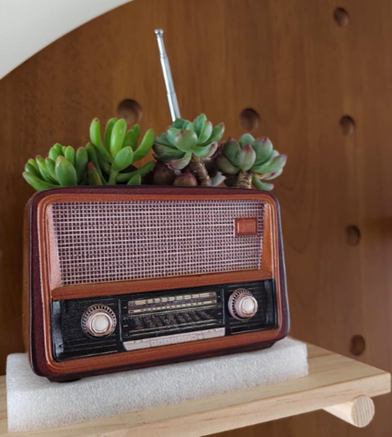 May include: A vintage-style radio planter with a brown wooden frame and a mesh speaker cover. The radio is filled with green succulents. The radio has two knobs and a silver antenna. The planter sits on a white foam base.