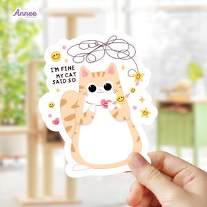 May include: A die-cut sticker featuring a cartoon orange tabby cat with large black eyes, holding a small pink heart. The text reads I'M FINE MY CAT SAID SO, accompanied by a dark brown scribble line, yellow stars, and small pink smiley face beads.