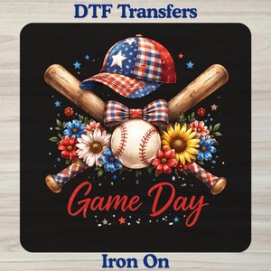 May include: A DTF transfer design featuring the text DTF Transfers at the top and Iron On at the bottom. The graphic shows a baseball, two crossed wooden bats, a plaid baseball cap, a plaid bow, and colorful flowers with the text Game Day.