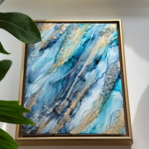 May include: Abstract painting in shades of blue, white, and gold with glitter accents. The painting is framed in a gold frame.