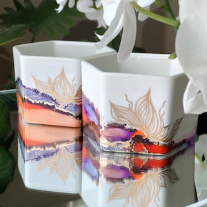 May include: Two white hexagonal ceramic candle holders with gold leaf and purple and orange abstract designs. The candle holders are sitting on a reflective surface.