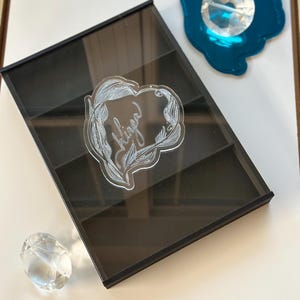 May include: A black acrylic jewelry display case with multiple compartments. A clear heart-shaped sticker with the name "Klinga" is on the front. A clear crystal and a teal-colored decorative piece are also present.